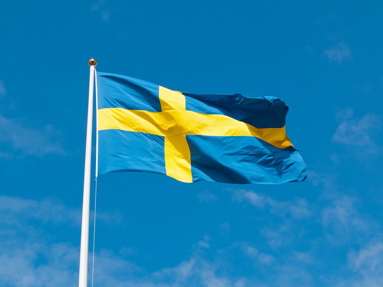 Sveriges flagga fladdrar i vind mot blå himmel med molnstrimmor